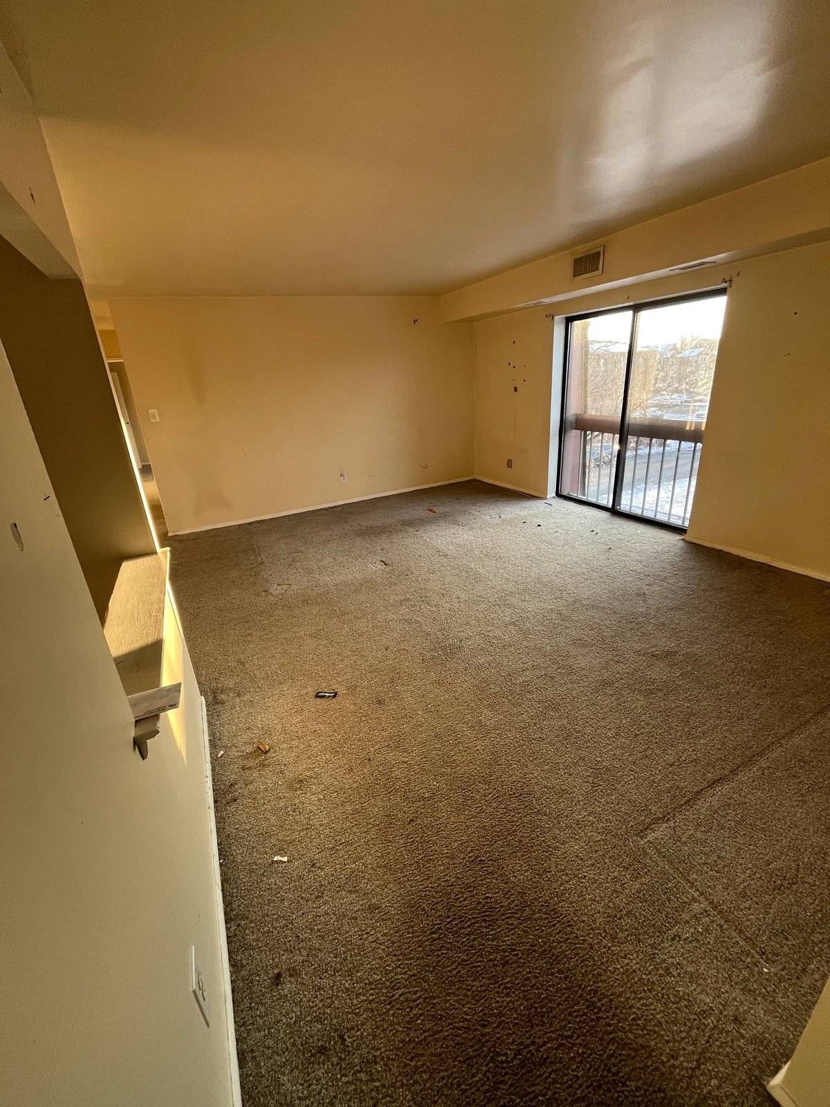 Empty apartment living room with tan walls, gray carpet flooring, and glass doors opening to a balcony with winter landscape view