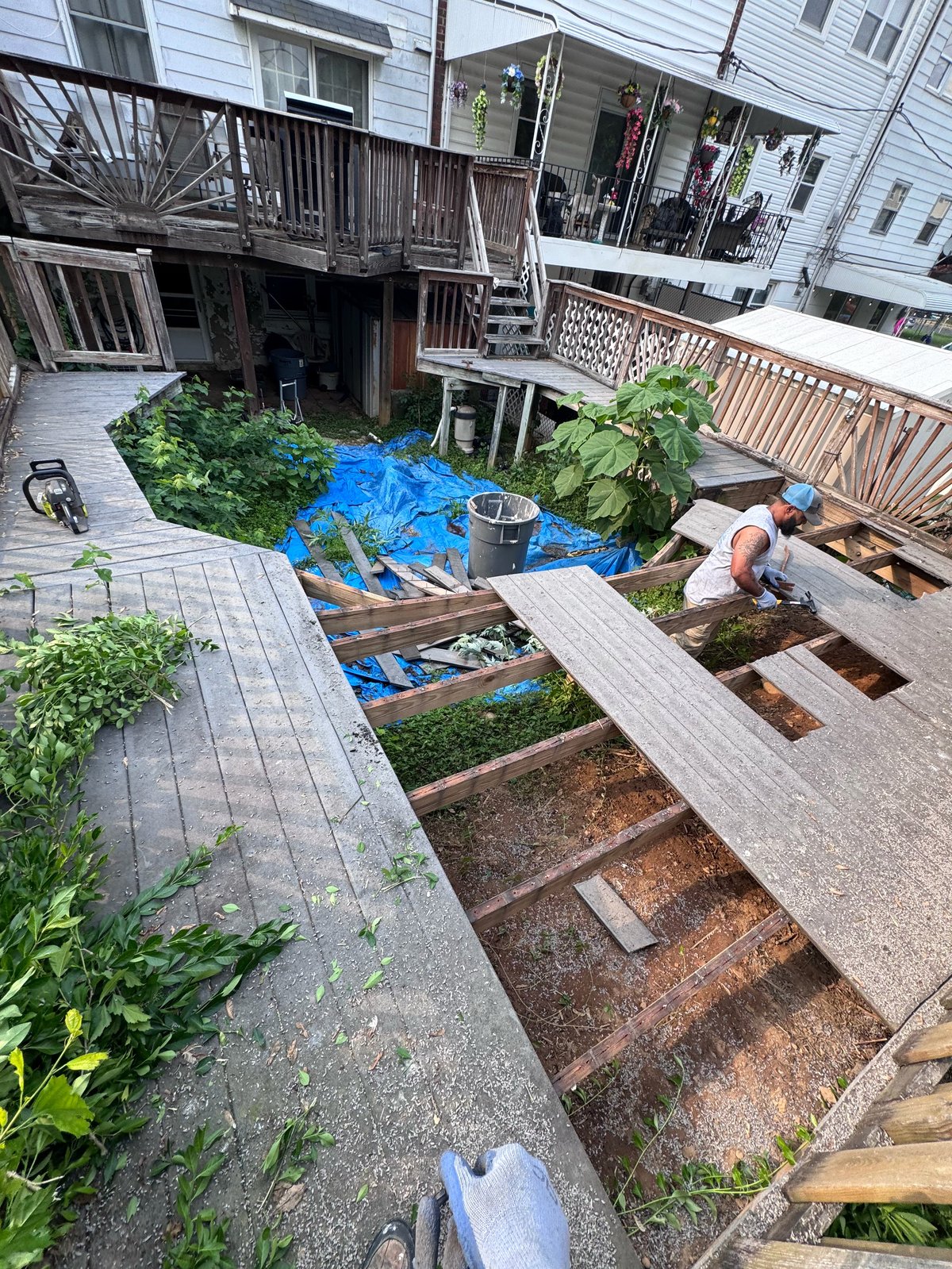 Overhead view of a residential backyard renovation in progress with wooden deck framing, blue tarps, and a worker building new outdoor living spaces