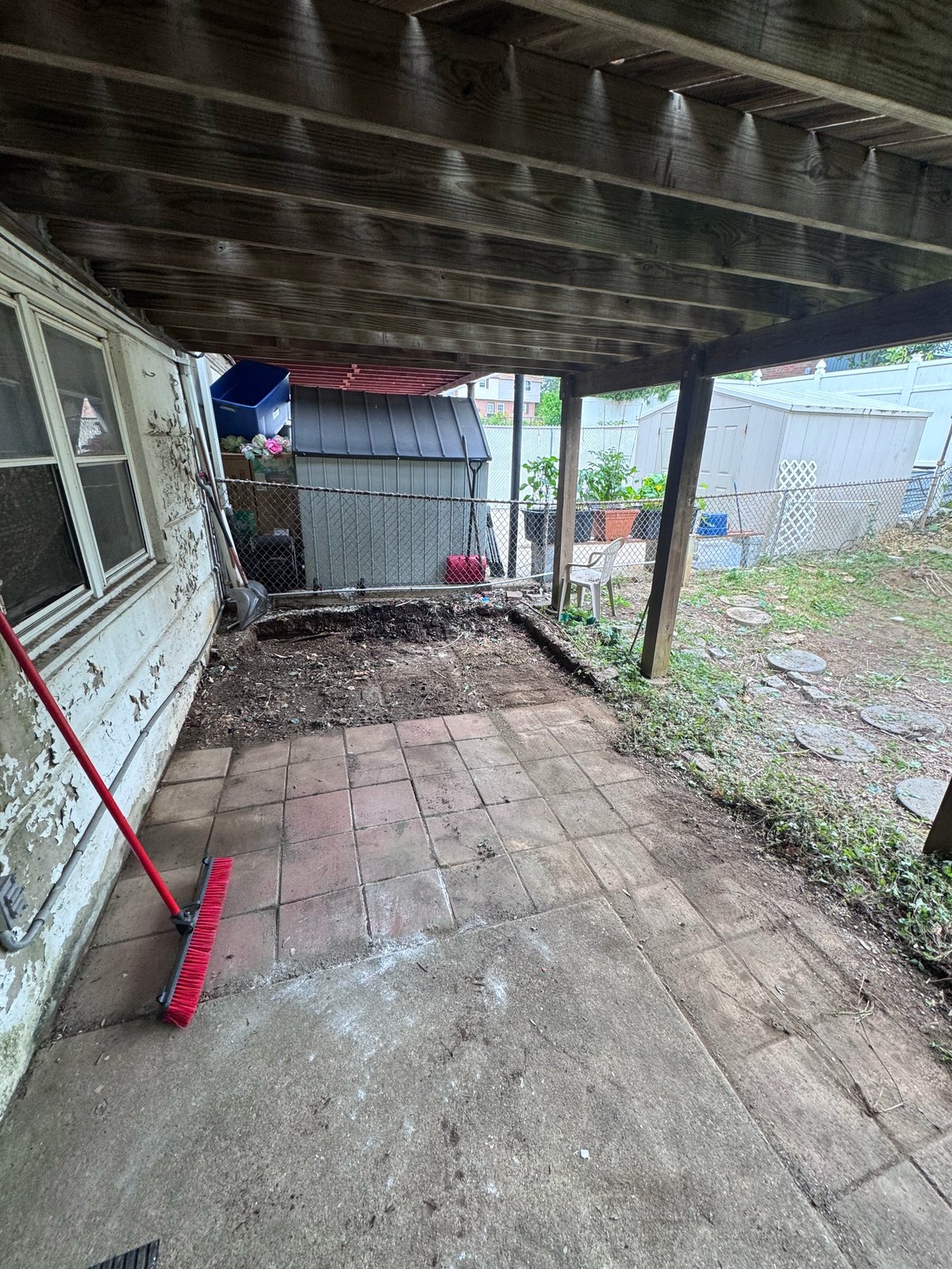 Covered patio area with brick floor, metal roof, and red rake, adjacent to residential structures and fenced yard